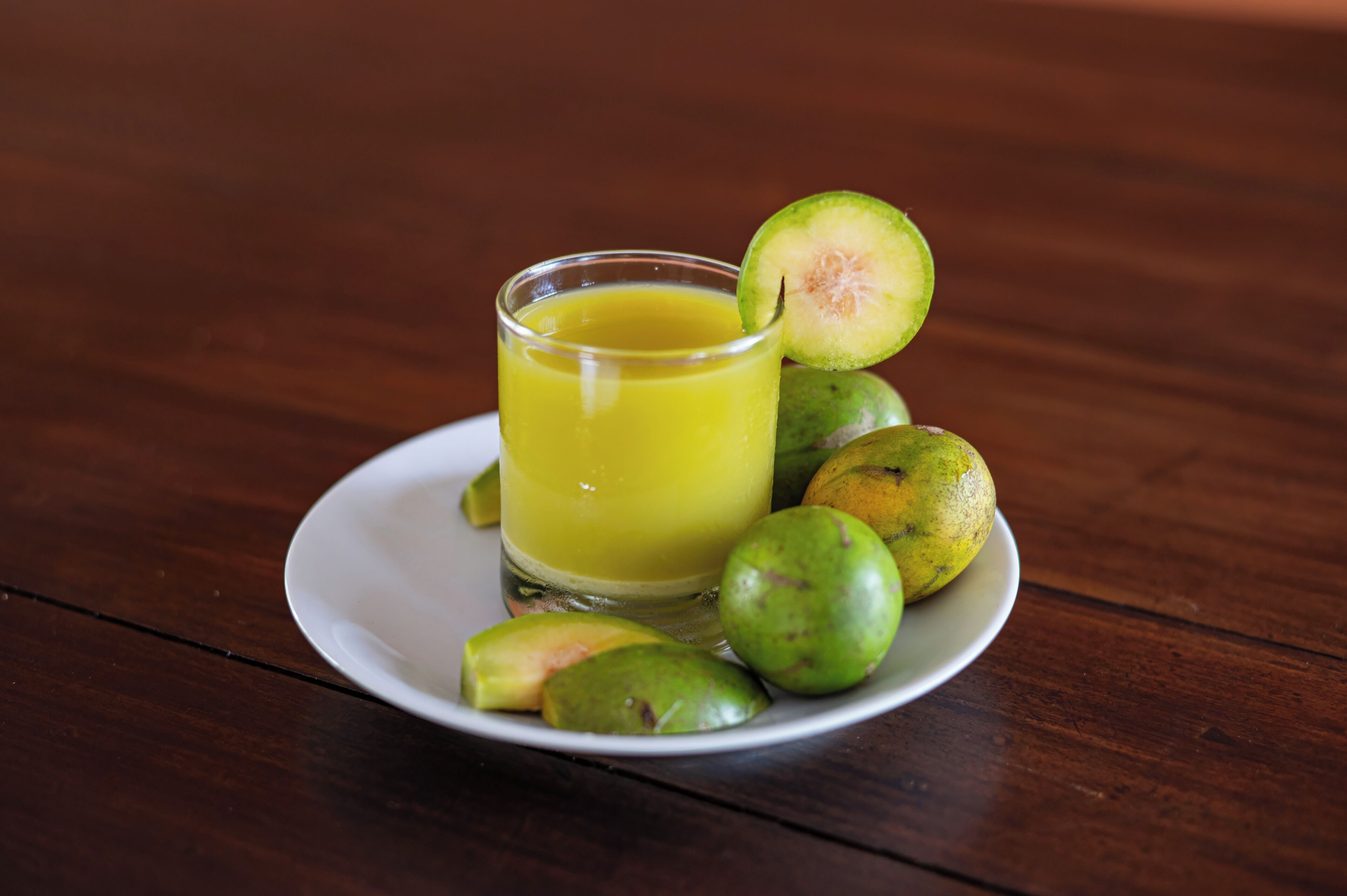 A glass of yellow juice on a white plate with several green fruits beside it.