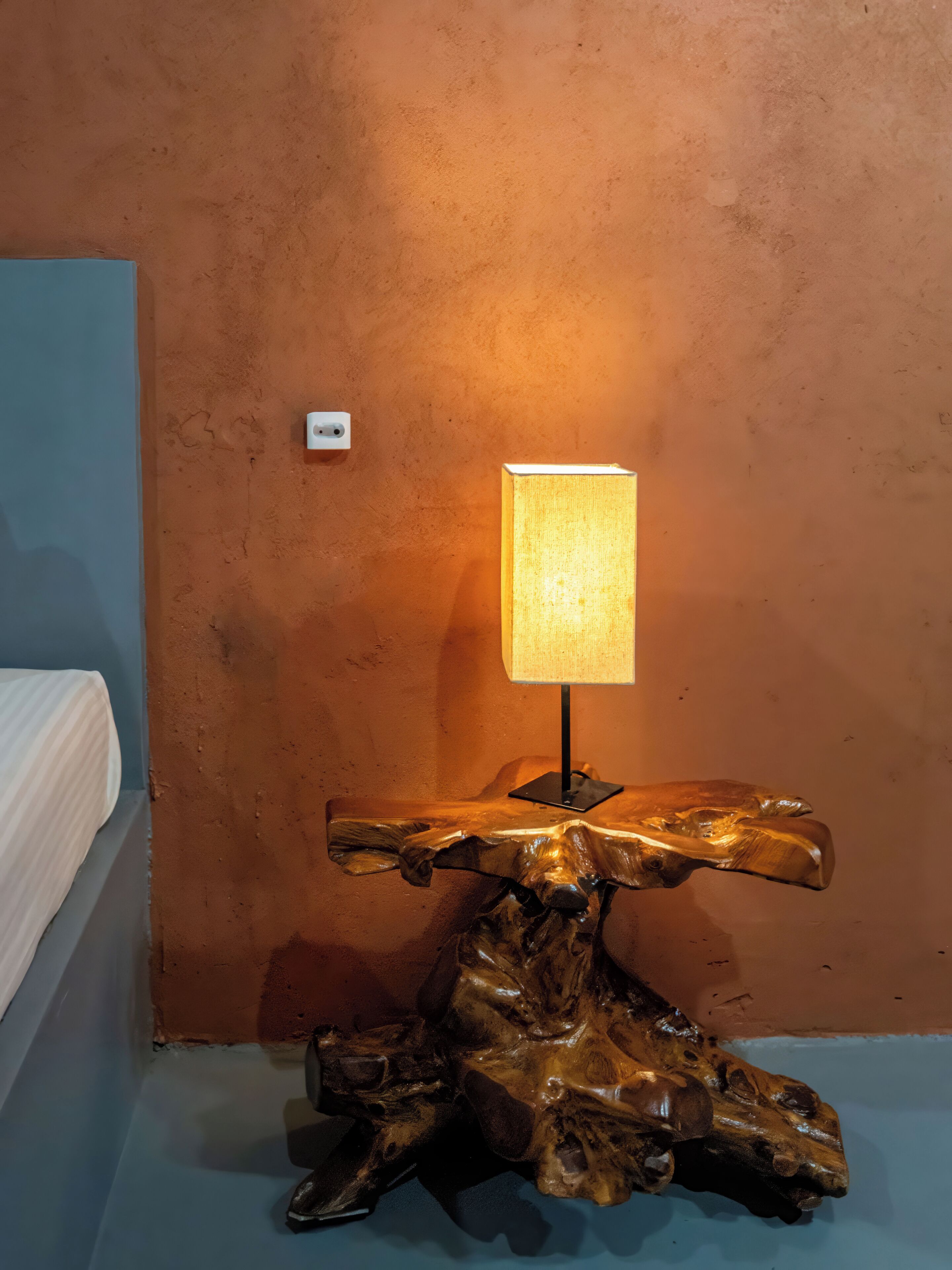 A wooden table with a lamp in a room with a textured orange wall and a blue floor.