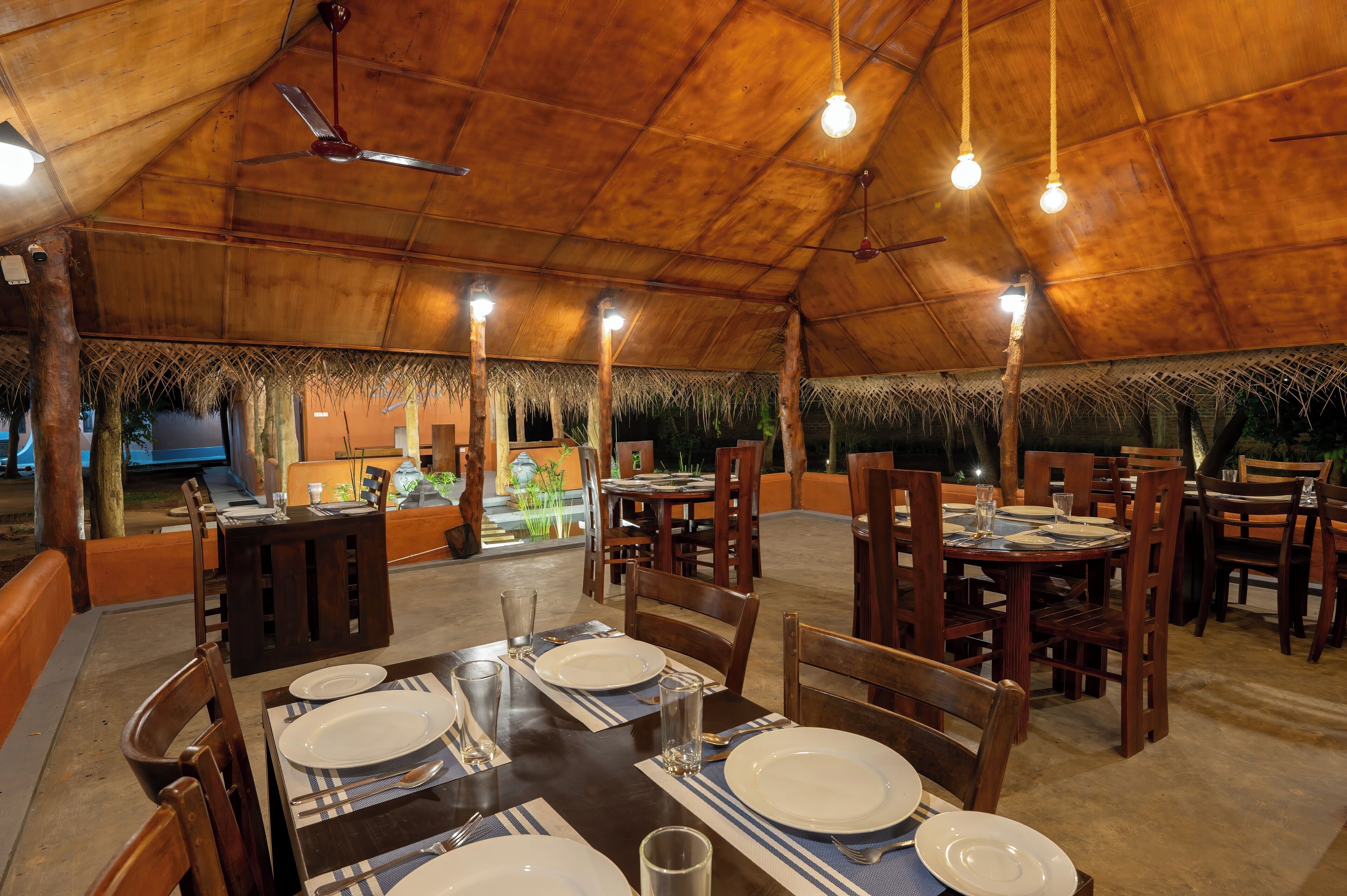 Un restaurante con mesas y sillas de madera, techo de paja y luces colgantes.