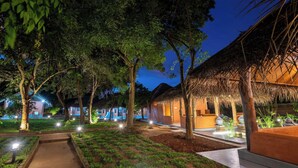 Un espace extérieur bien éclairé, doté de constructions à toits de chaume, entouré d’arbres et agrémenté de chemins illuminés.