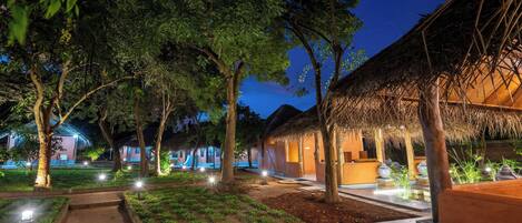 A well-lit outdoor area with thatched-roof structures, surrounded by trees and illuminated pathways.