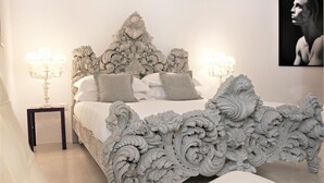 A bedroom with an ornate bed frame, two bedside lamps, and a framed portrait on the wall. - Reggia del Saraceno – Luxury House in the Heart of Ortigia (Siracusa)