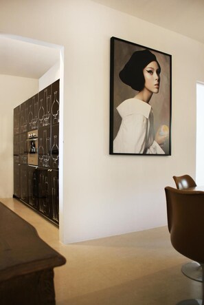 A modern kitchen with a wine rack, a dining area with a chair, and a framed portrait on the wall. - Reggia del Saraceno – Luxury House in the Heart of Ortigia (Siracusa)