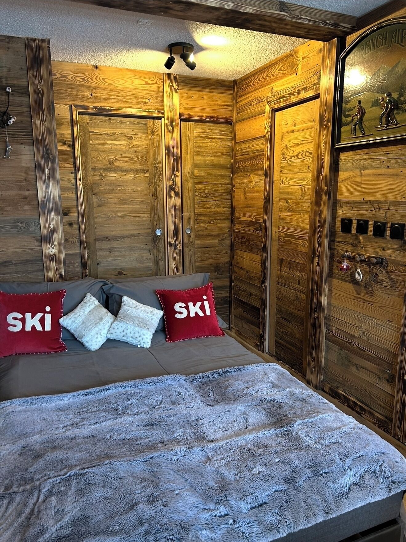 A cozy room with wooden walls, a bed with a grey blanket, and red pillows with the word "SKI".