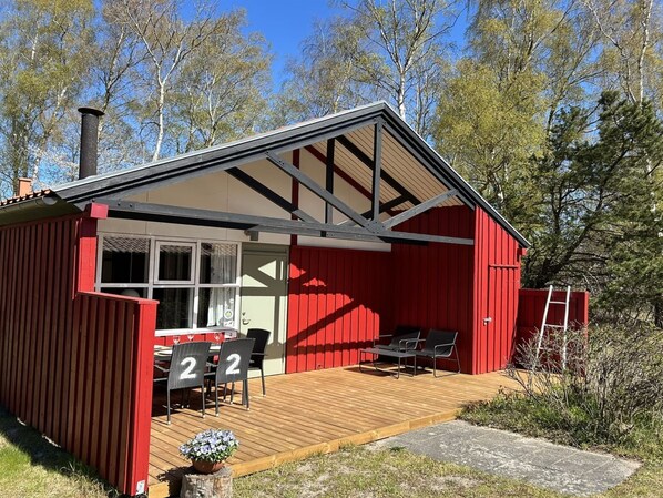 Una cabaña roja con una terraza de madera, una mesa y dos sillas, y una chimenea.