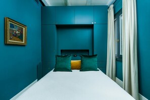 Ein Schlafzimmer mit türkisfarbenen Wänden, einem Bett mit weißer Bettwäsche, zwei türkisfarbenen Kissen und einem gerahmten Bild an der Wand.