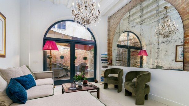 A modern living room with a sofa, two armchairs, a coffee table, and a chandelier. There are large windows with arched tops, a brick wall, and a view of an outdoor patio. - The Secret Garden of Venice Sanctuary: A Peaceful Getaway (Venezia)