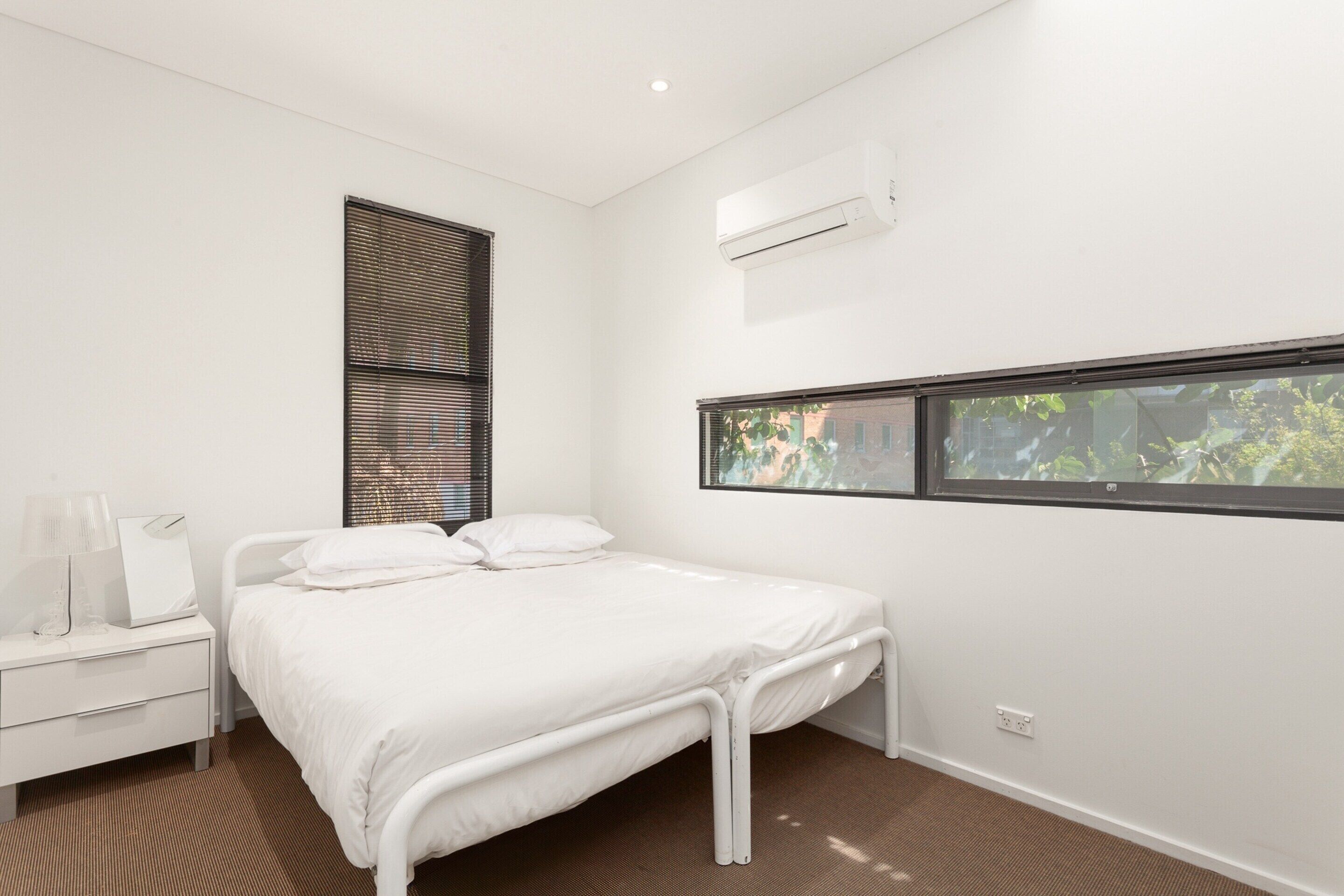 Une chambre moderne avec un lit simple, une table de chevet, une fenêtre avec des stores et un appareil de climatisation.