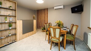 Un comedor con sillas de madera, una mesa, un televisor montado en la pared y una estantería de madera con plantas.