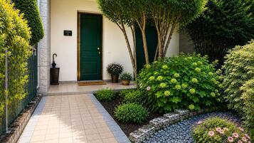 Un giardino curato con un vialetto piastrellato che conduce a una porta verde, circondato da arbusti diversi e una pianta in vaso.