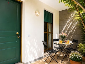 Una habitació amb una porta verda, una finestra amb persianes verdes, taula i cadires, i una planta en test.