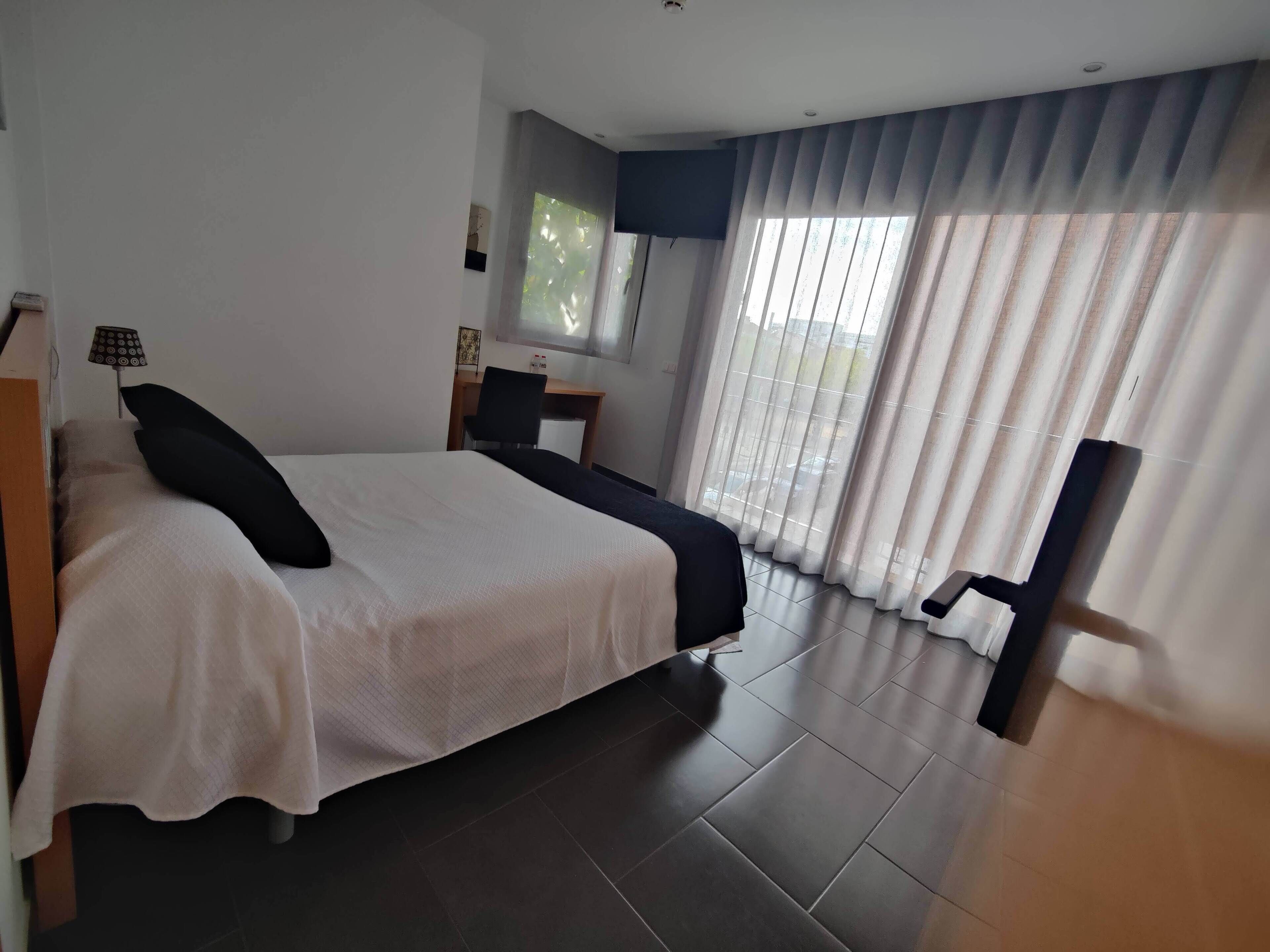 Een moderne hotelkamer met een groot bed, een bureau en een stoel. Er is een raam met gordijnen, een flatscreen televisie en een lamp op het bureau.