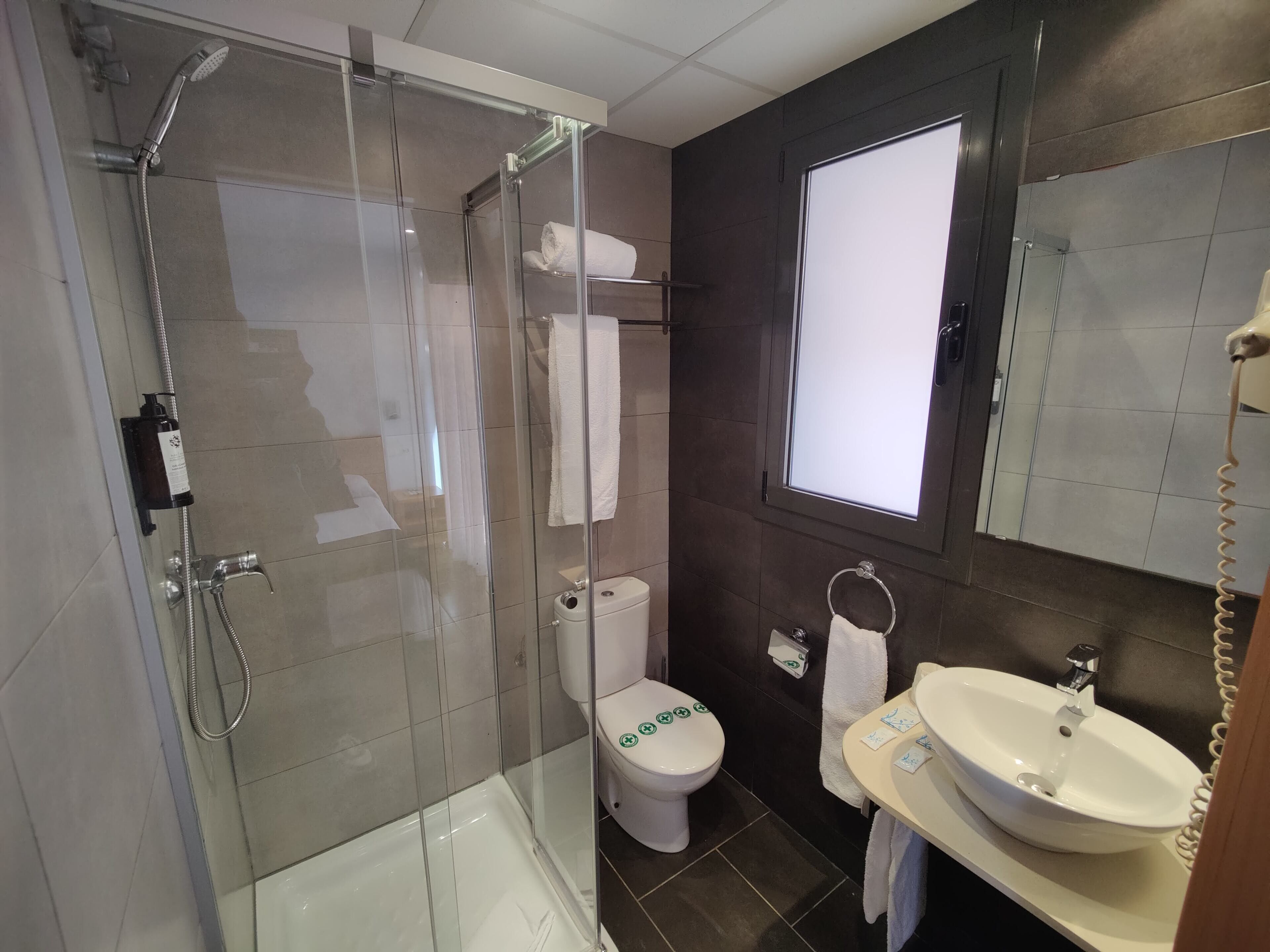 Een moderne badkamer met een douche met glazen wand, een toilet met een groene sticker, een wastafel en een spiegel.