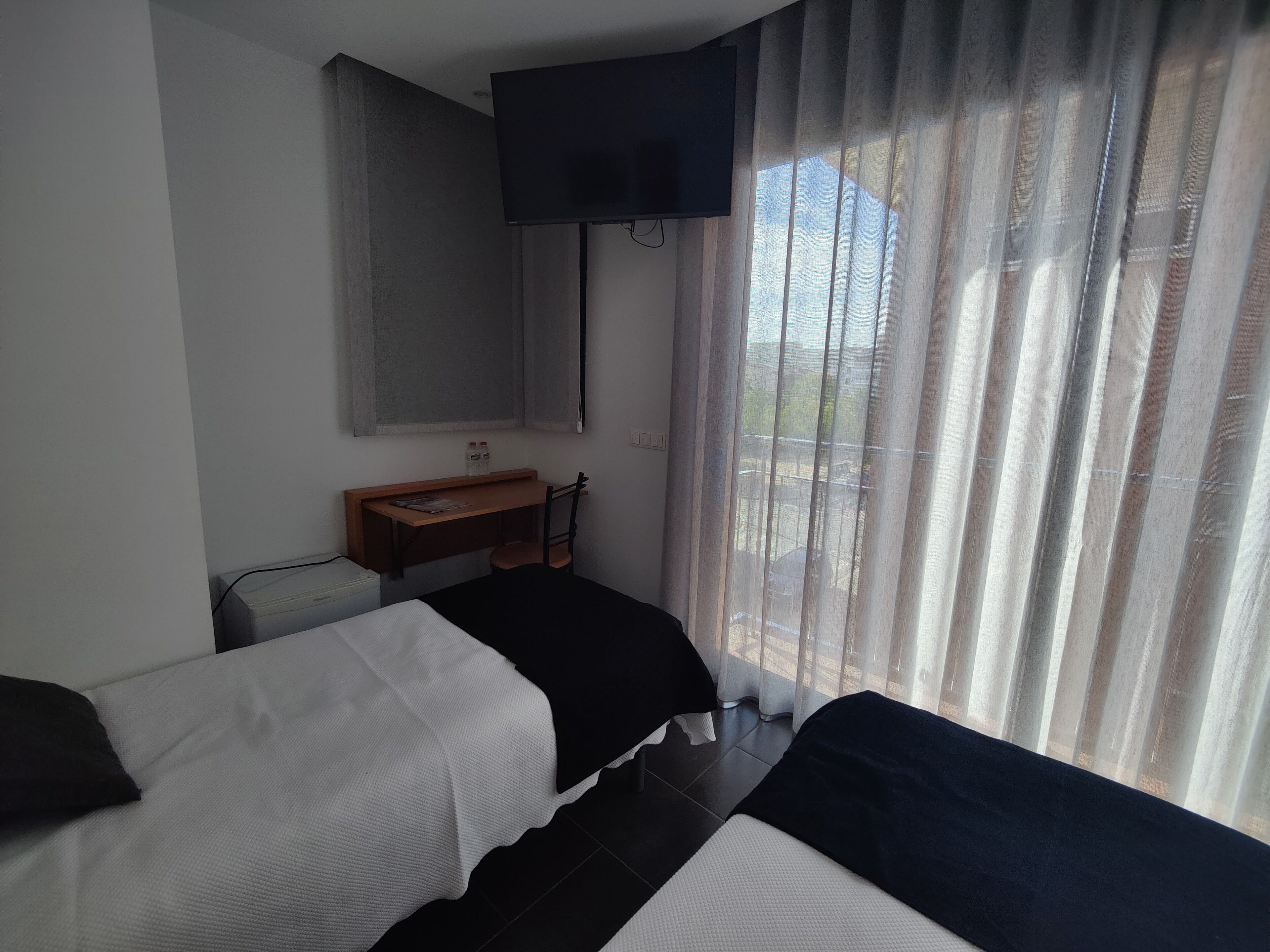 Habitación de hotel con dos camas, un escritorio, televisión montada en la pared y una ventana con cortinas translúcidas.