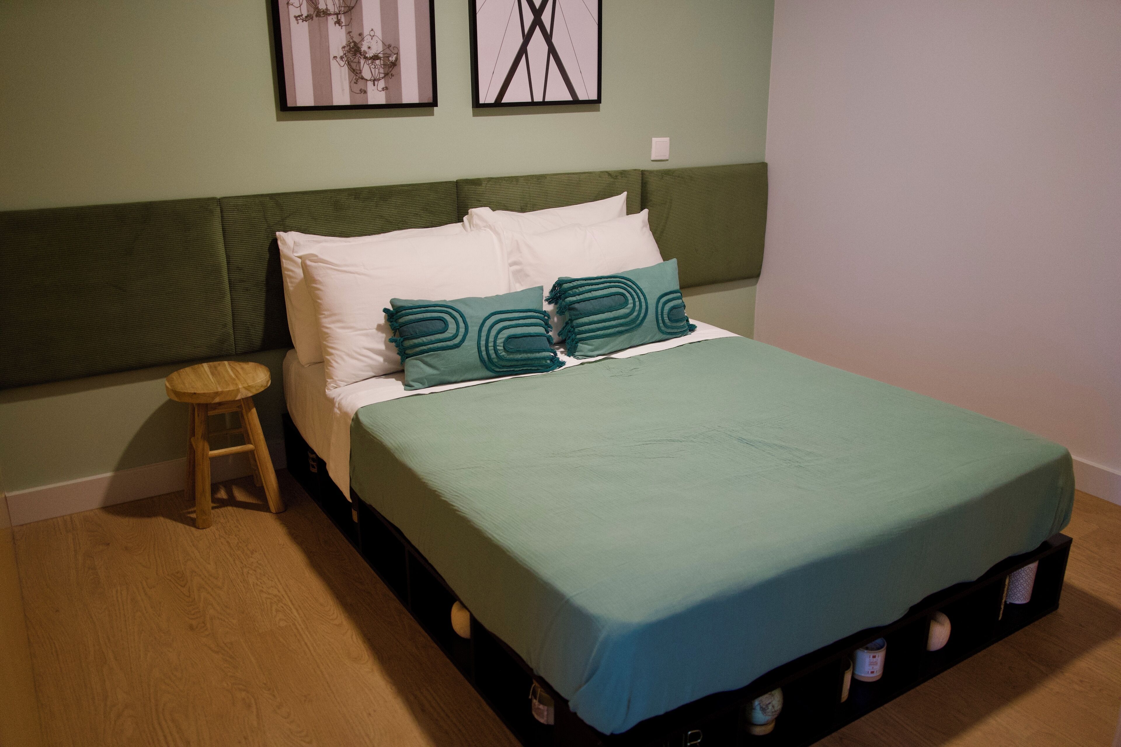 Una cama con colcha verde y almohadas turquesa, un taburete de madera y dos cuadros en la pared.