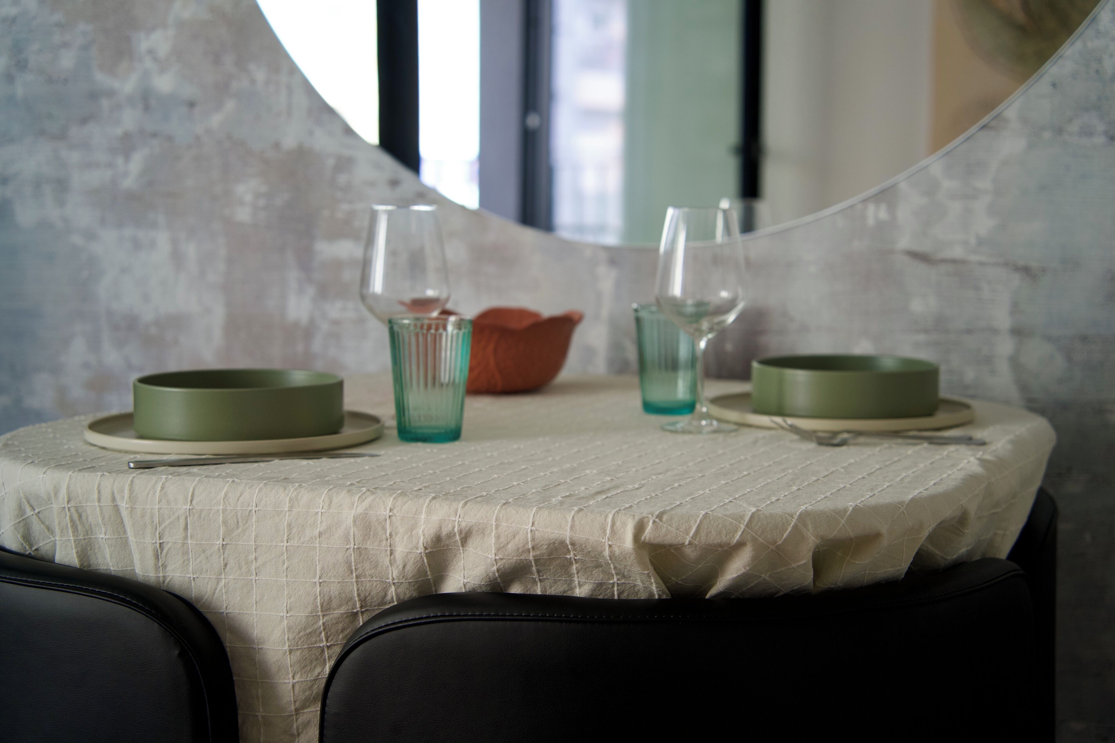 Una mesa de comedor con mantel blanco, dos platos verdes, dos vasos y un bol naranja pequeño.