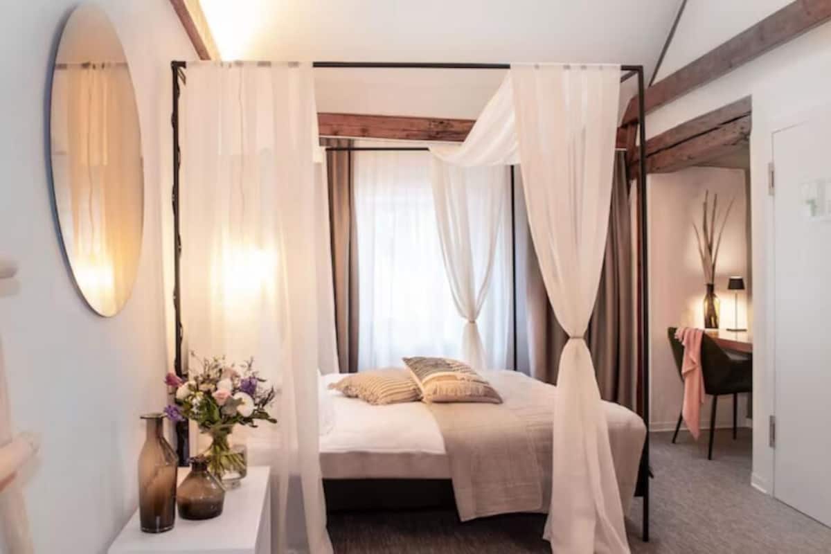 A bedroom with a four-poster bed, a bedside table with a vase of flowers, and a large mirror on the wall.