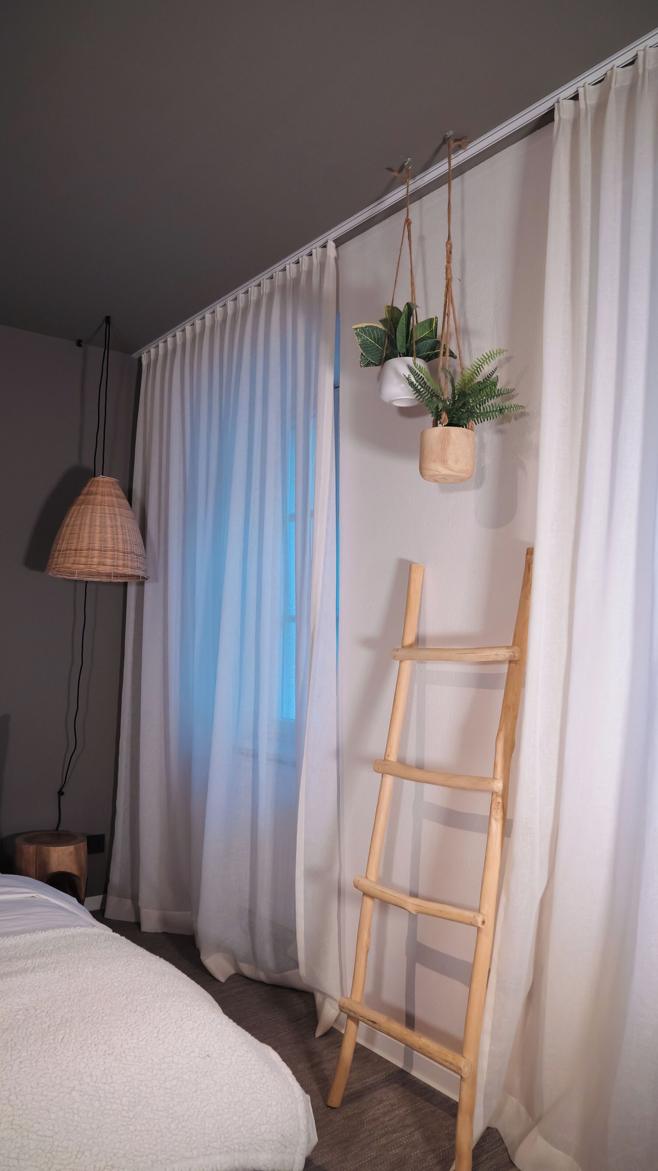 A bedroom with a bed, a hanging plant, a wooden ladder, and a window with sheer curtains.