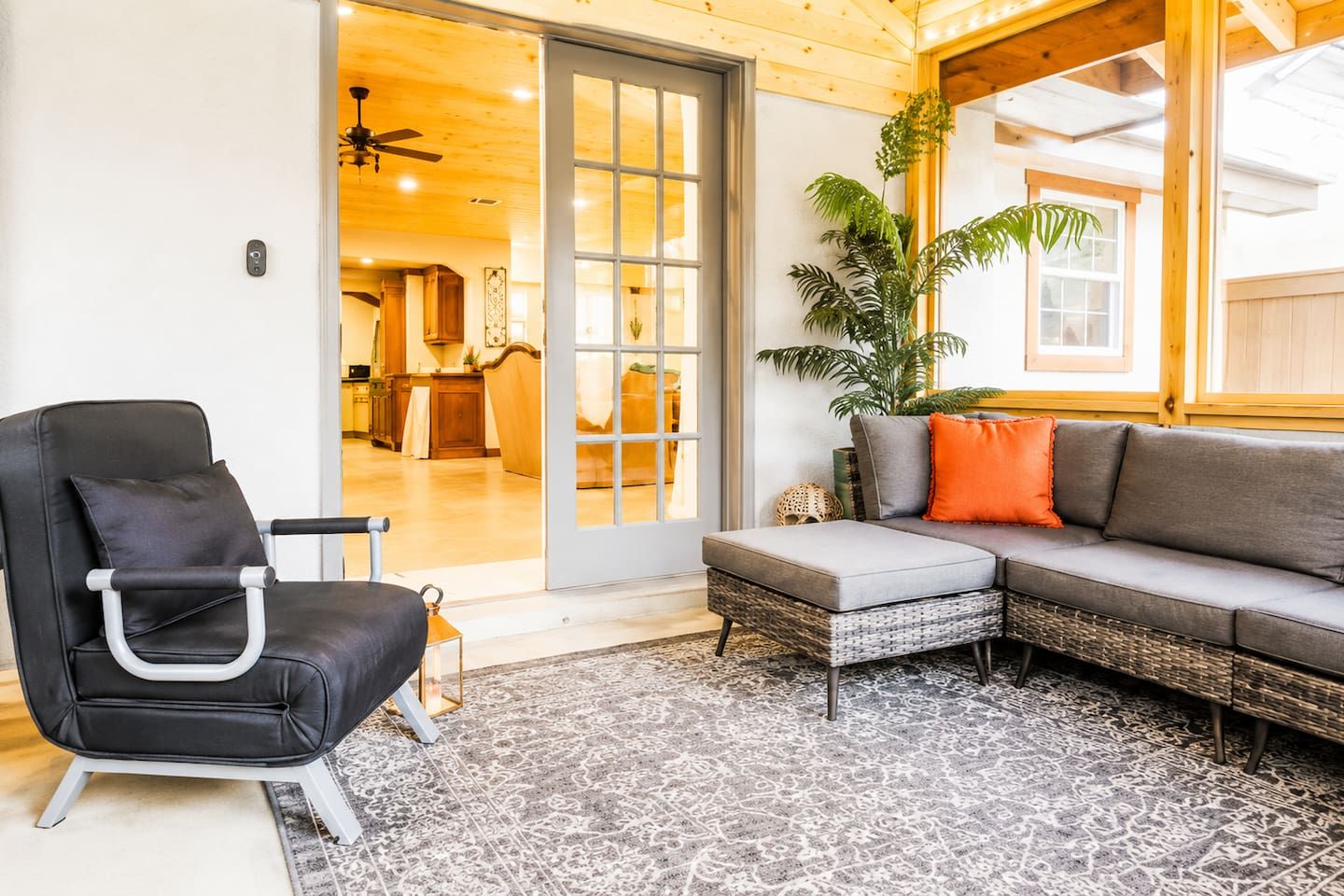 A room with a black chair, a grey sofa, a patterned rug, and a large window with a view into the kitchen.