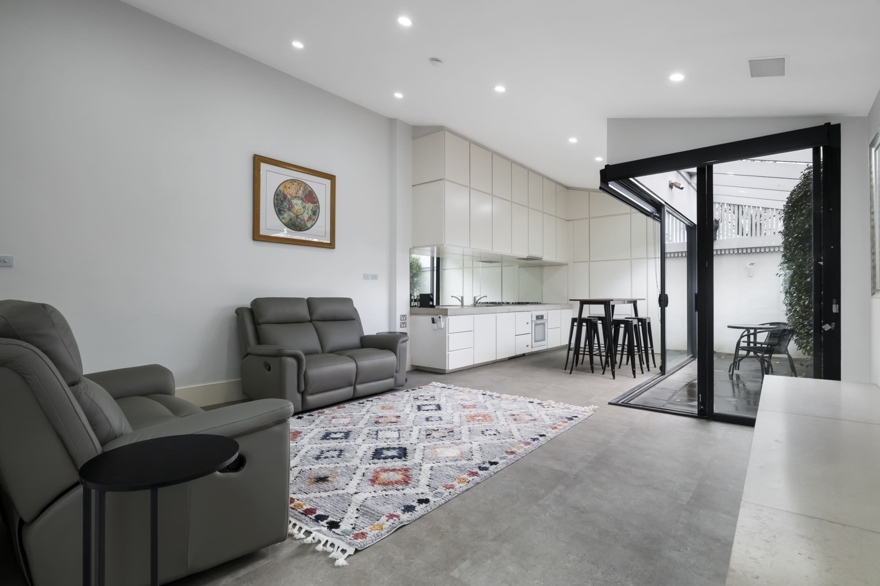 A modern living room with a grey sofa, a coffee table, and a patterned rug. There is a kitchen area with white cabinets and a dining space with stools.