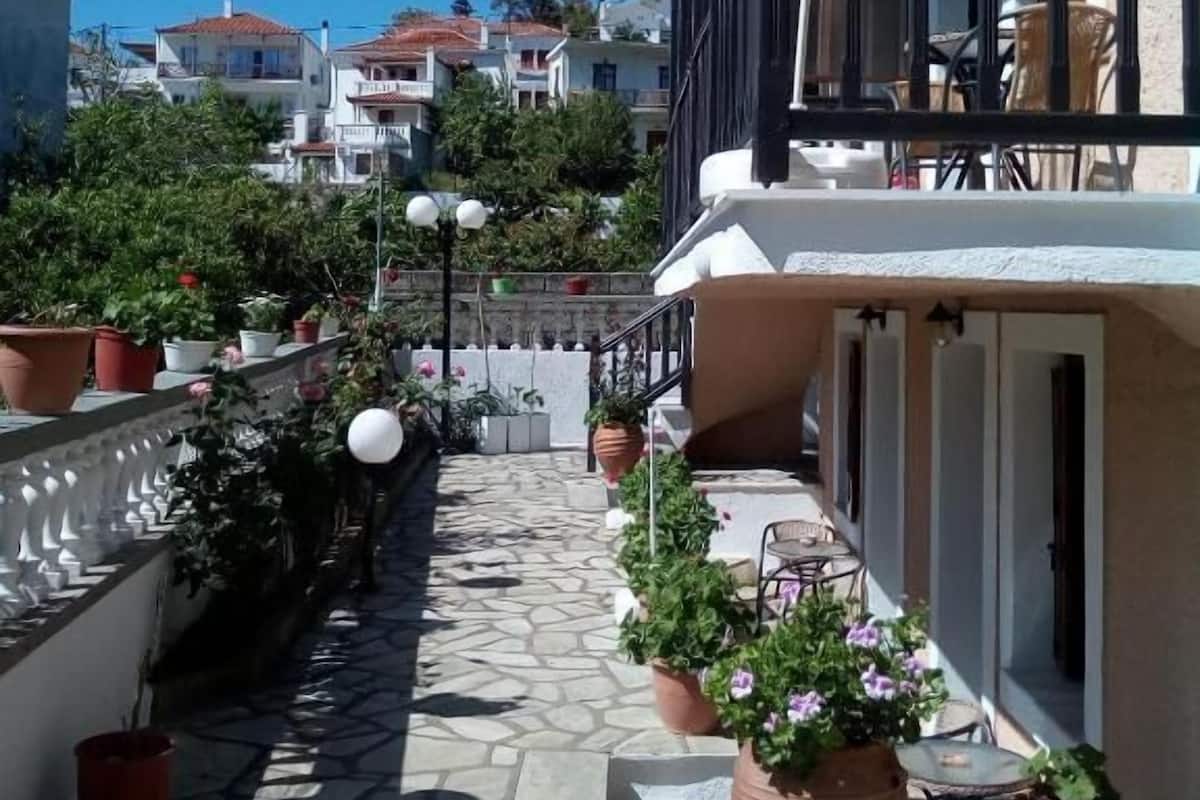 A narrow stone-paved pathway leading to a building with a balcony, potted plants, and outdoor furniture.