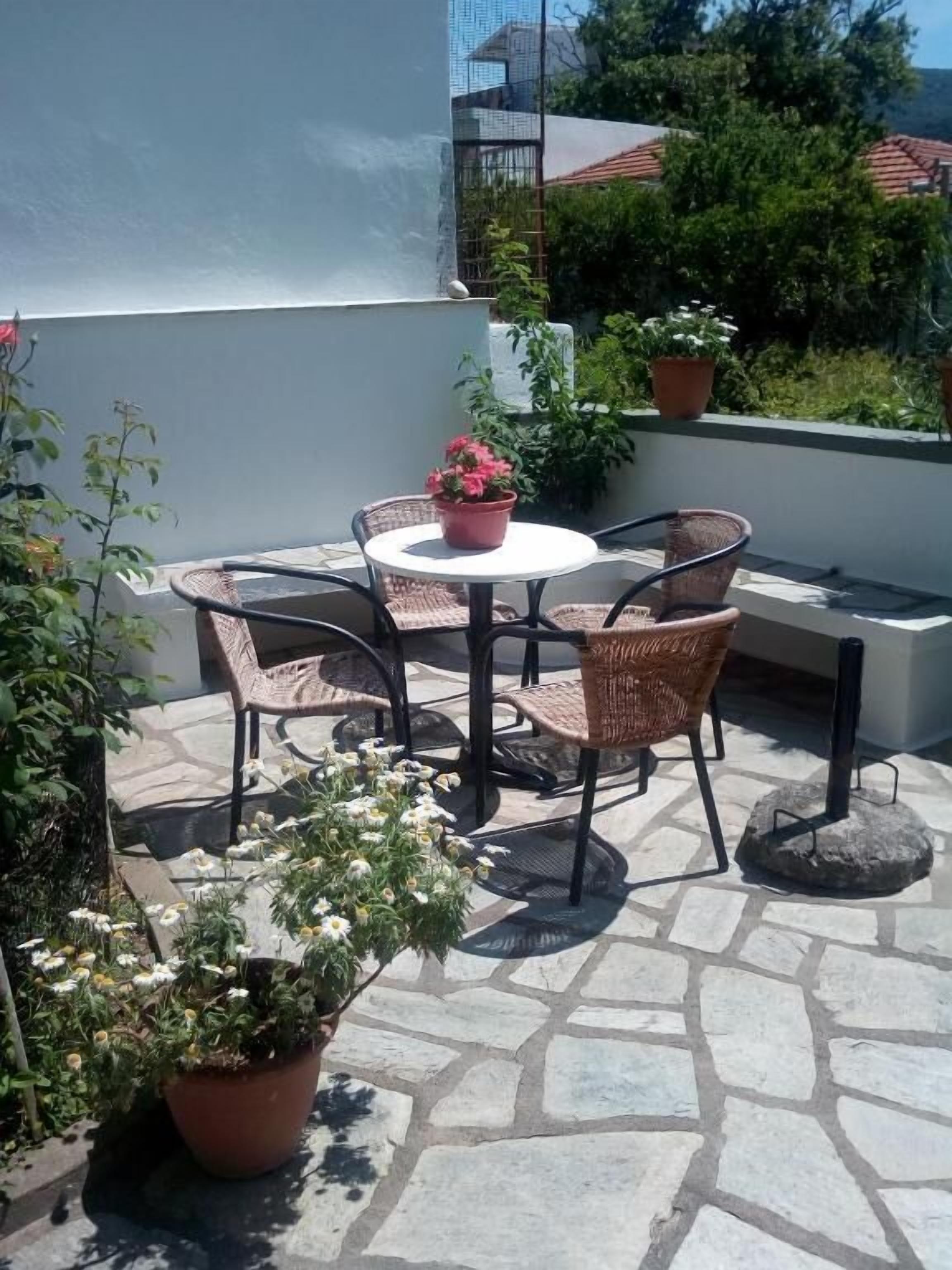 Een terras buiten met een ronde tafel en rieten stoelen, omringd door plantenpotten en een stenen vloer.