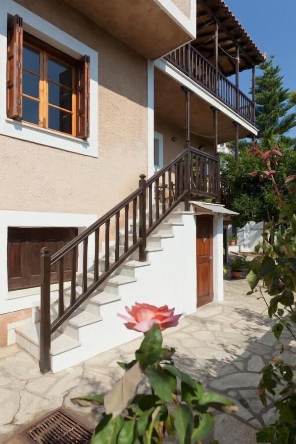 A two-story building with a stone-paved entrance and a wooden staircase with a metal railing. - Sofia's Garden by Booking Kottas (Skopelos)