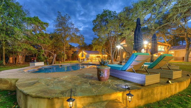 Un espace extérieur bien éclairé autour d’une piscine, avec des chaises longues et des parasols, le tout entouré d’arbres et bordé d’une terrasse en pierre.