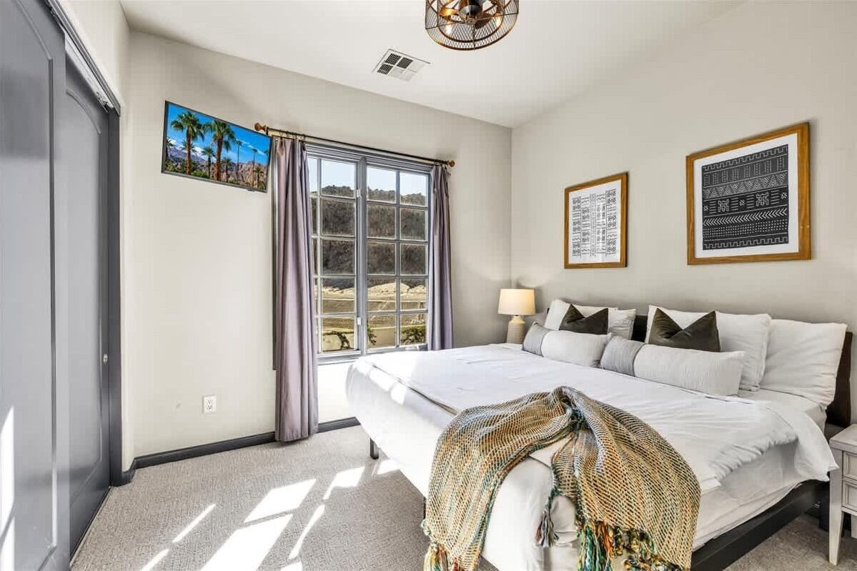 Un dormitorio con una cama grande, una ventana con cortinas, una lámpara de pared y obras de arte enmarcadas.