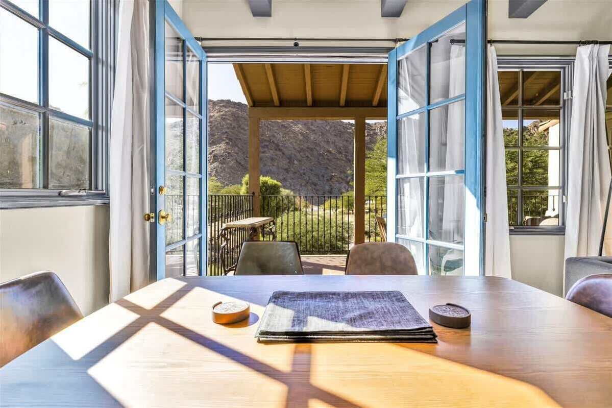 Una habitación con una mesa de madera, sillas y vistas a un paisaje montañoso a través de puertas abiertas.