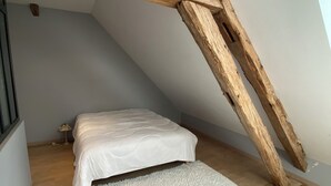 Un dormitorio con techo inclinado, vigas de madera, una cama, alfombra blanca y una mesita de noche pequeña.