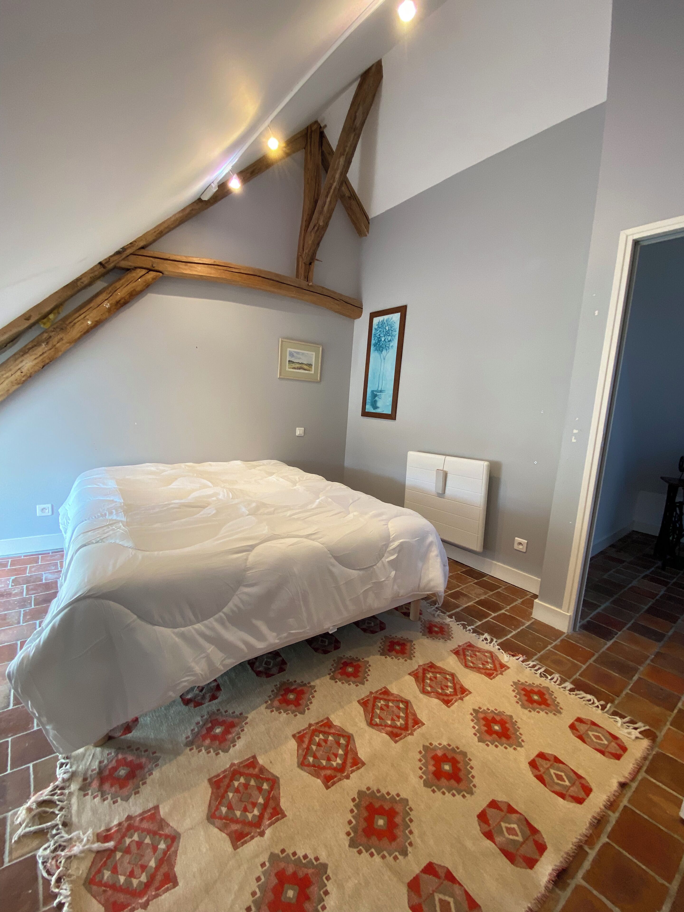 Une chambre avec un lit, un tapis à motifs, un radiateur et une poutre en bois au plafond.
