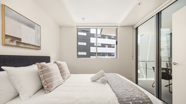Ein modernes Schlafzimmer mit einem großen Bett, Blick auf ein Gebäude durch ein Fenster und einer Schiebeglastür zum Balkon.
