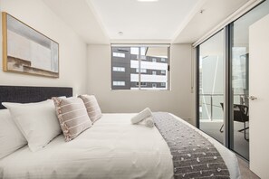 Un dormitorio moderno con una cama grande, vistas de un edificio a través de una ventana y una puerta corrediza de vidrio a un balcón.