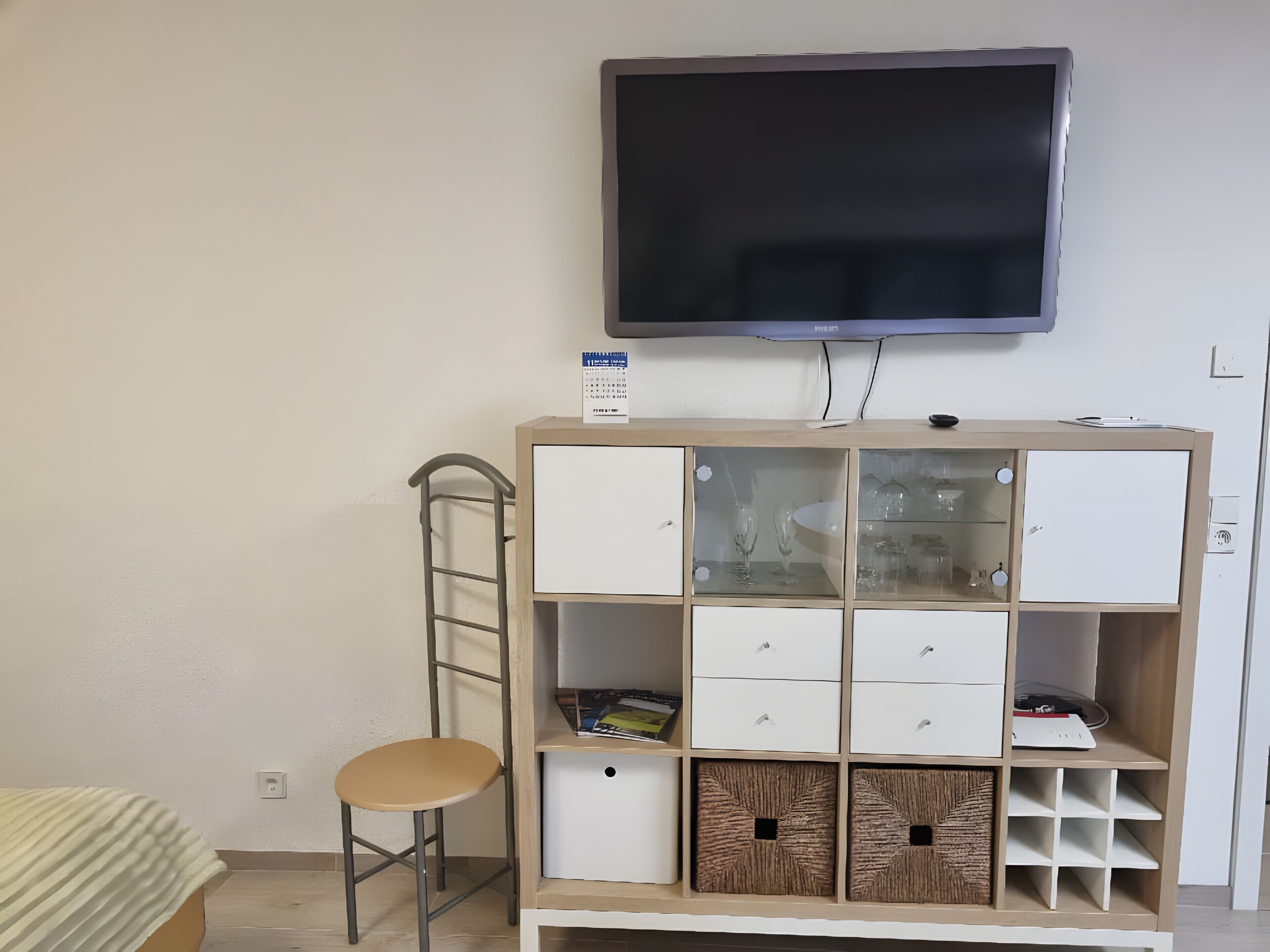 Ein Zimmer mit an der Wand montiertem Fernseher, einem Holzkasten mit Schubladen, einem Hocker und einem Bett mit gestreifter Bettdecke.