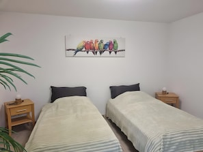 A room with two beds, a wall-mounted painting of colorful birds, a bedside table with a lamp, and a potted plant.