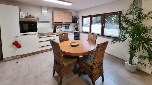 Une cuisine moderne avec des meubles blancs, un four encastré et un coin repas comprenant des chaises en osier et une table ronde.