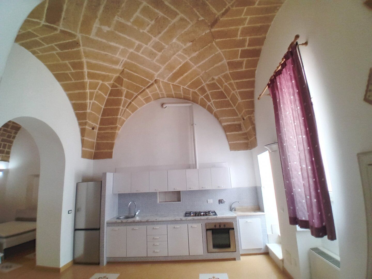 A compact kitchen with a brick ceiling, white cabinets, a built-in oven, and a window with purple curtains.