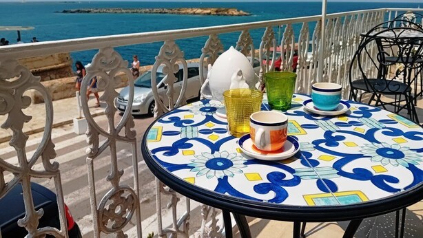 A round table with a blue and white patterned tablecloth, a white ceramic pitcher, and two glass tumblers on a balcony overlooking the sea. - Dimora San Felice - Il Mandorlo (Gallipoli)