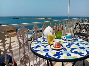 Una mesa redonda con mantel estampado en azul y blanco, una jarrón de cerámica blanca y dos vasos de vidrio en un balcón con vistas al mar.