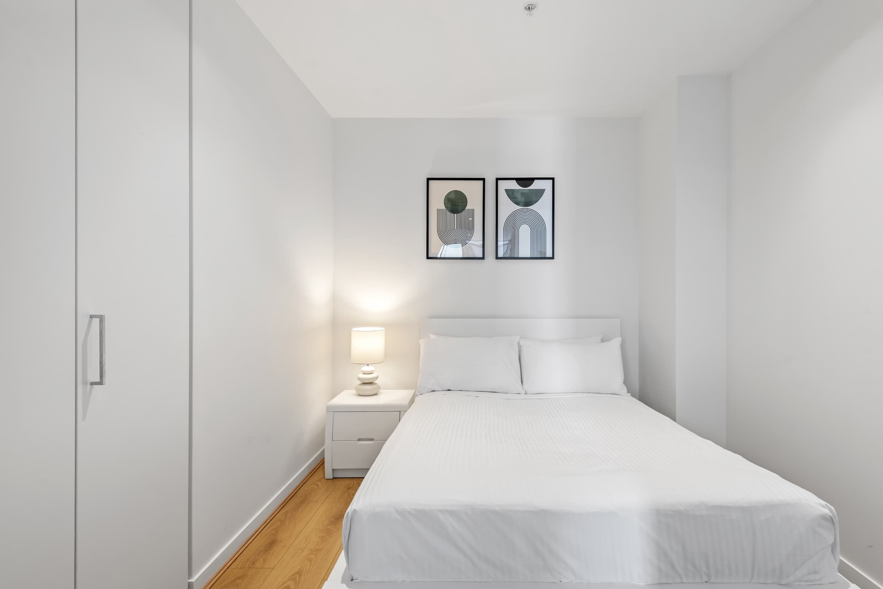A minimalist bedroom with a bed, nightstand, and two framed artworks on the wall.
