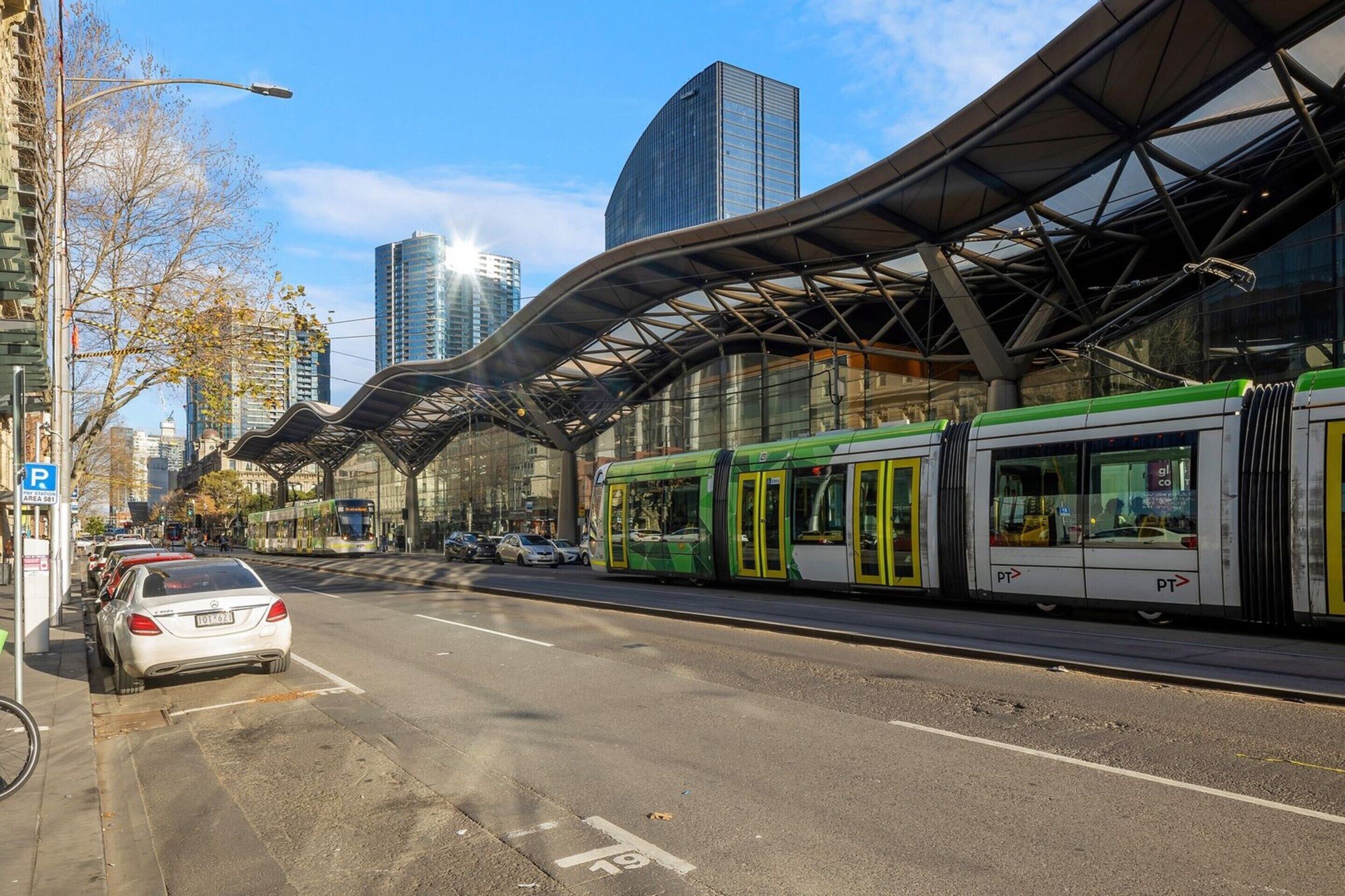 Een moderne stadstraat met een tramlijn, een overdekte voetgangerspassage en hoge gebouwen op de achtergrond.
