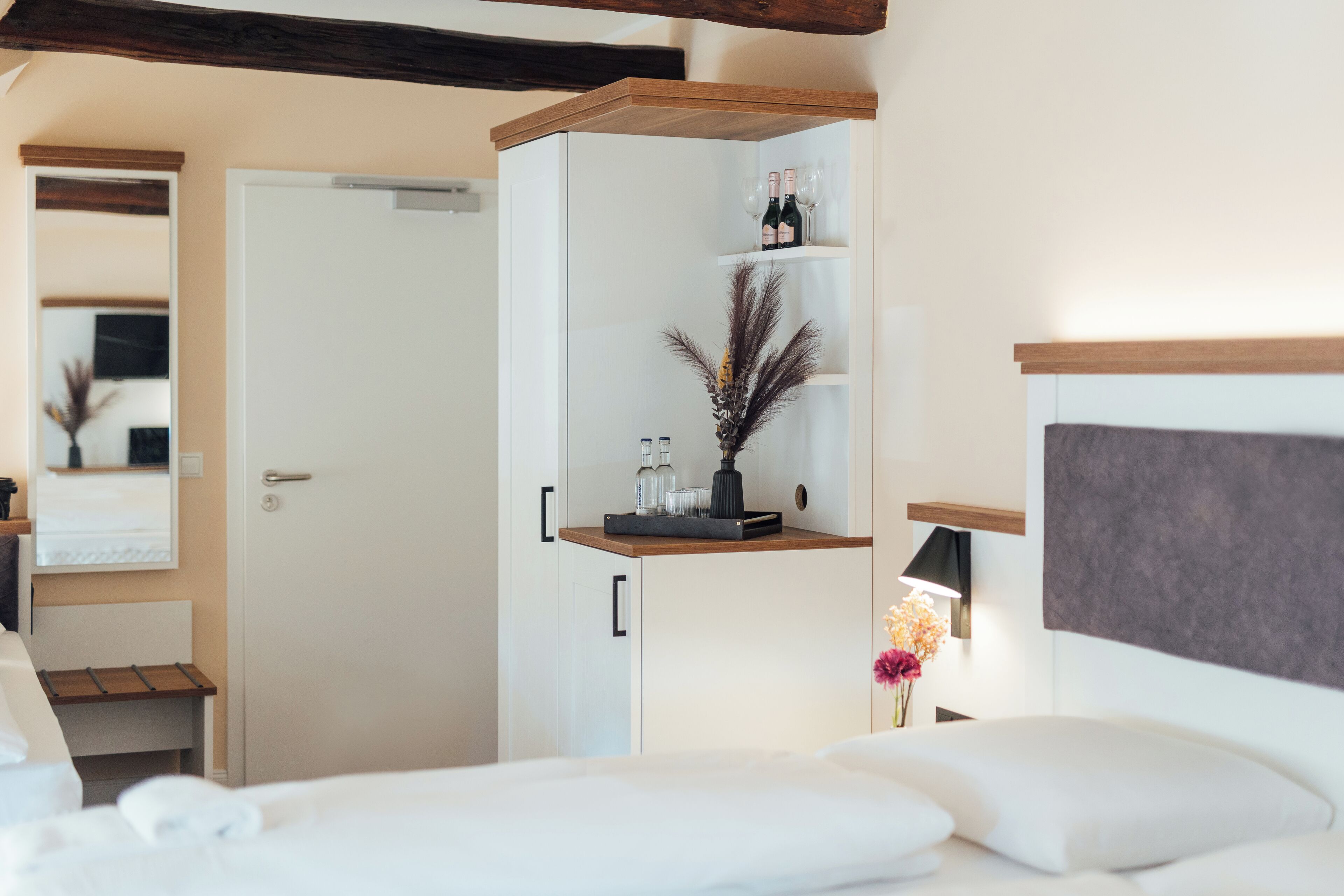 A hotel room with a bed, a wardrobe, a shelf with bottles and glasses, and a nightstand with a lamp and flowers.