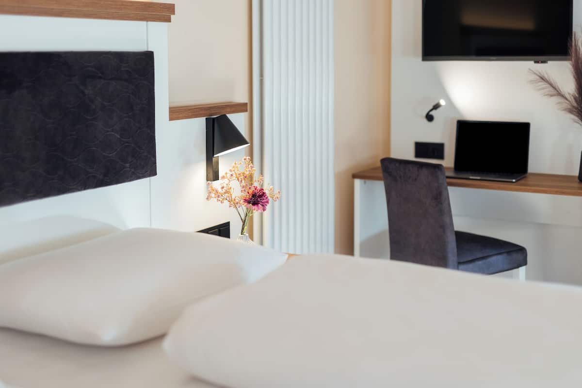 Une chambre d’hôtel moderne équipée d’un lit, d’un bureau avec un ordinateur portable, d’une télévision et d’un vase rempli de fleurs.