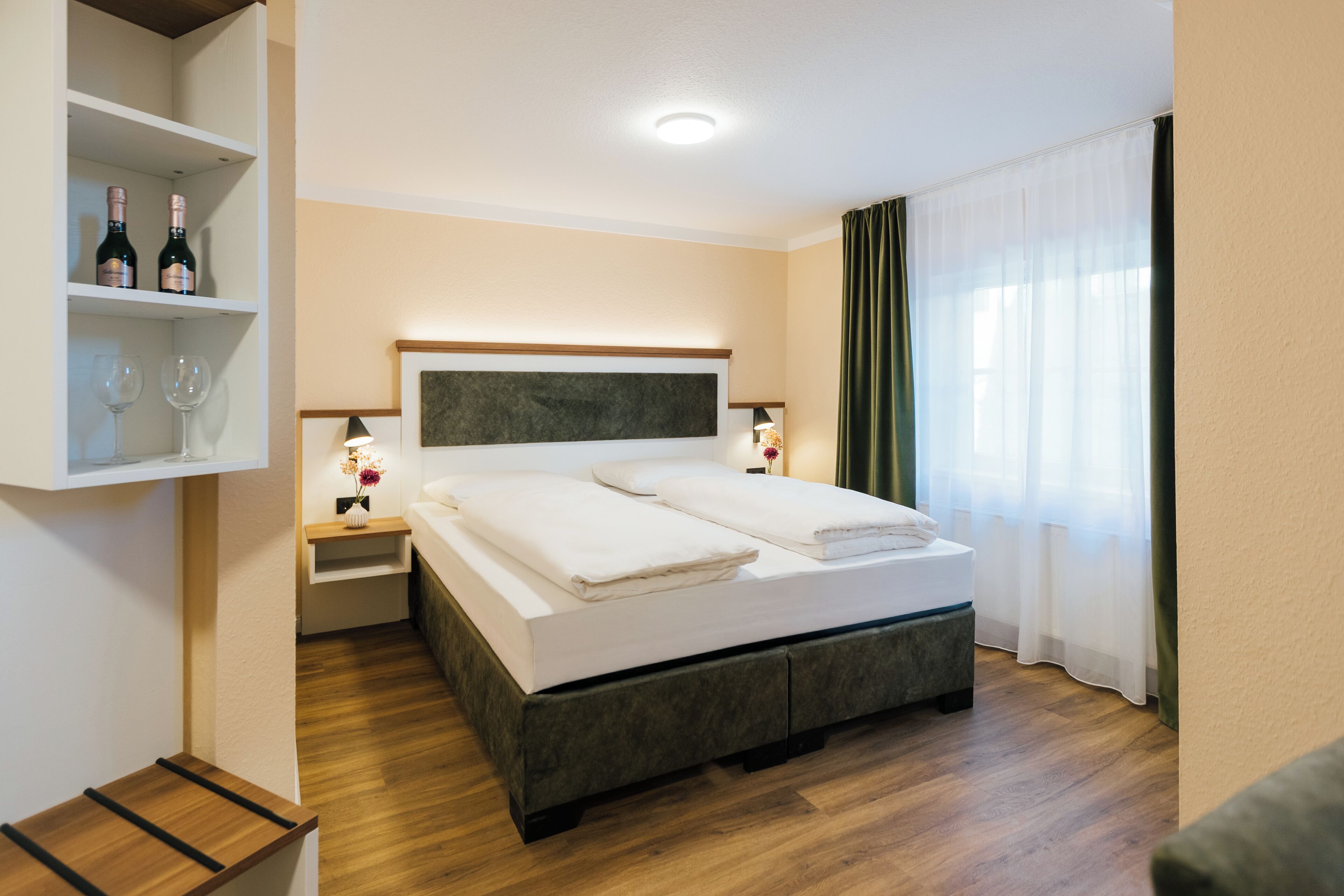A hotel room with a bed, two bedside tables, a shelf with wine bottles and glasses, and a window with curtains.