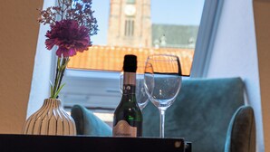 Une fenêtre donnant sur le clocher d’une église, un vase rempli de fleurs, une bouteille et deux verres à vin posés sur une table.