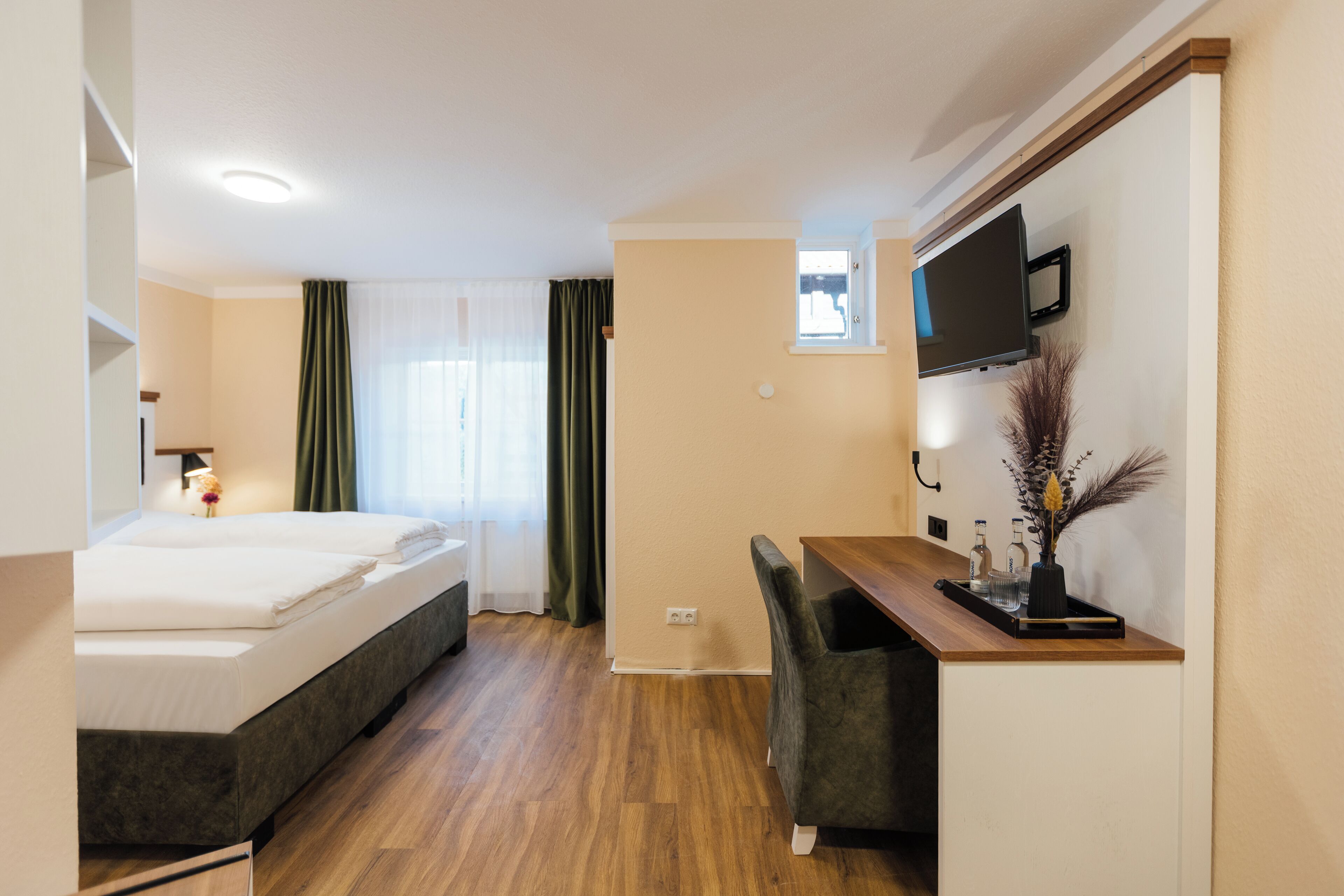 Habitación de hotel con cama, escritorio, silla, televisor y ventana con cortinas.
