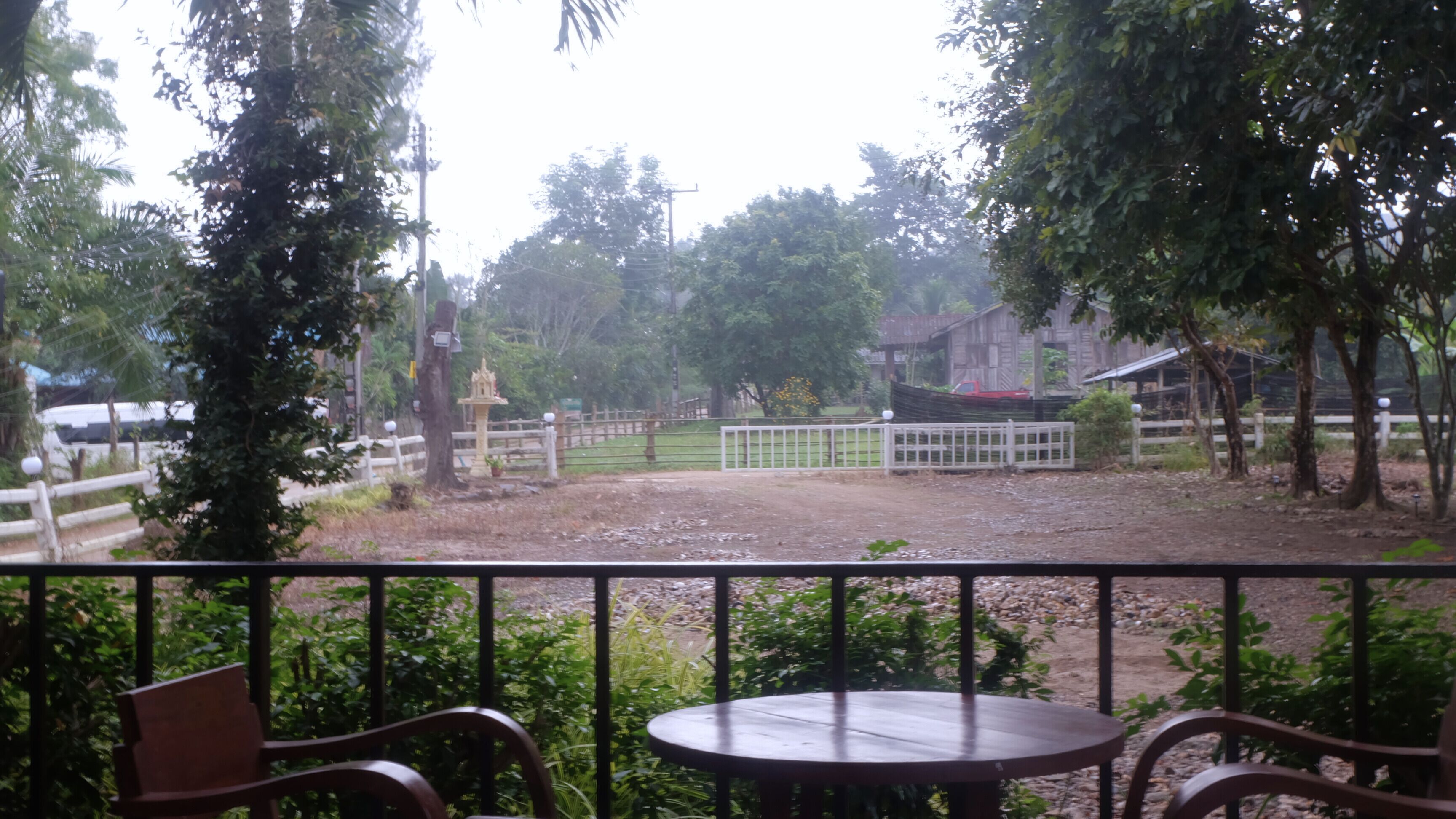 A view from a balcony with a table and chairs, overlooking a fenced area with trees and a small structure.