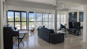 A spacious living room with a dark sofa, a glass coffee table, a flat-screen TV, and a piano. Large windows offer a view of the outdoors. - Bright, quiet loft within walking distance - Exceptional location (Le Gosier)