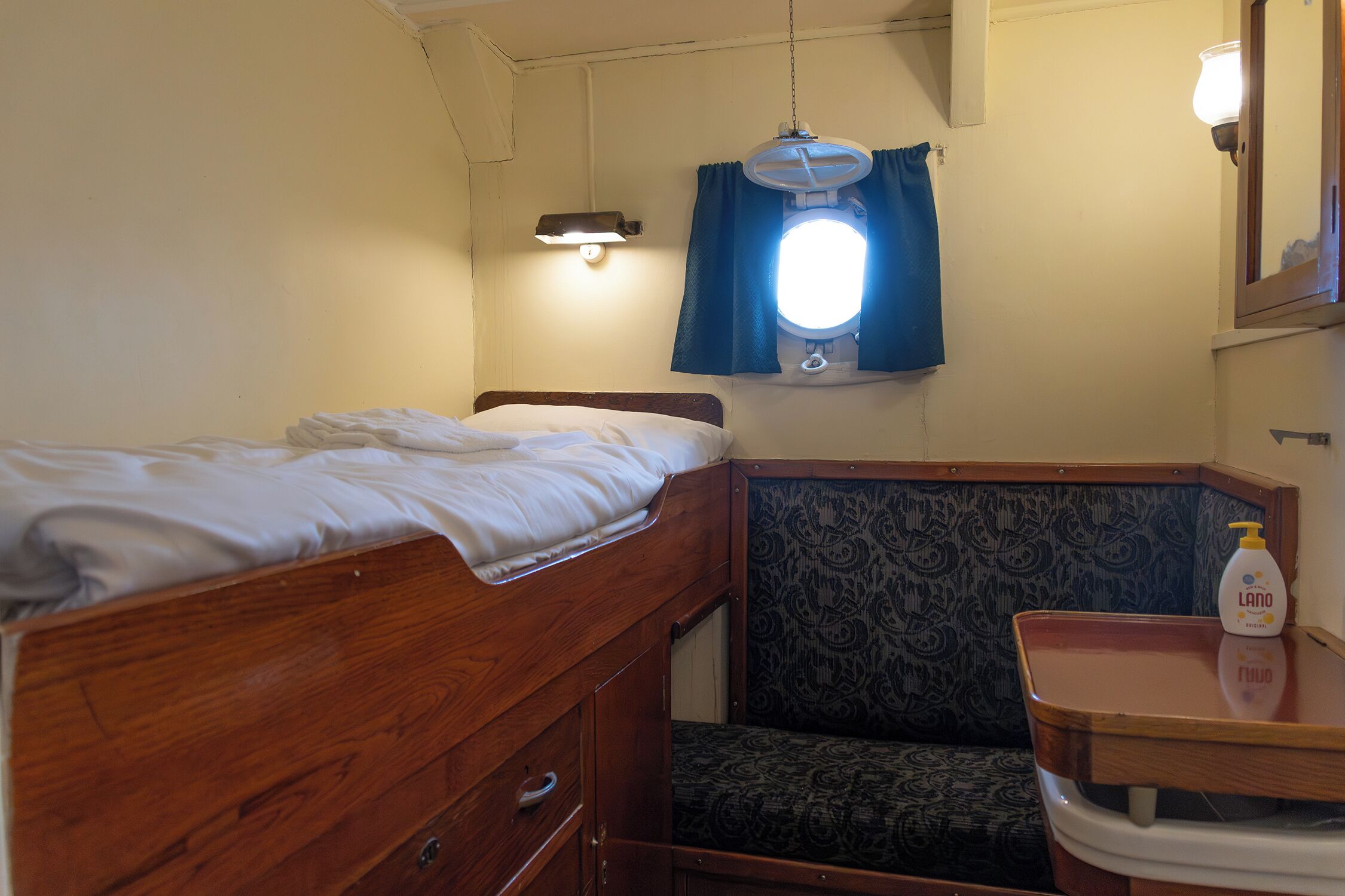 Een kleine, compacte kamer met een stapelbed, een stoel, een klein tafeltje met een fles en een wandlamp.